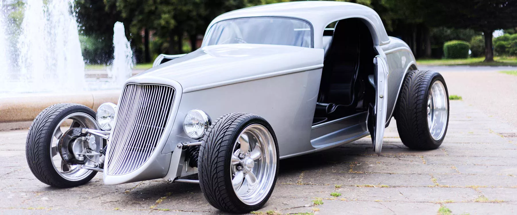 Silver Ford Model B hot rod with its door open in front of fountains