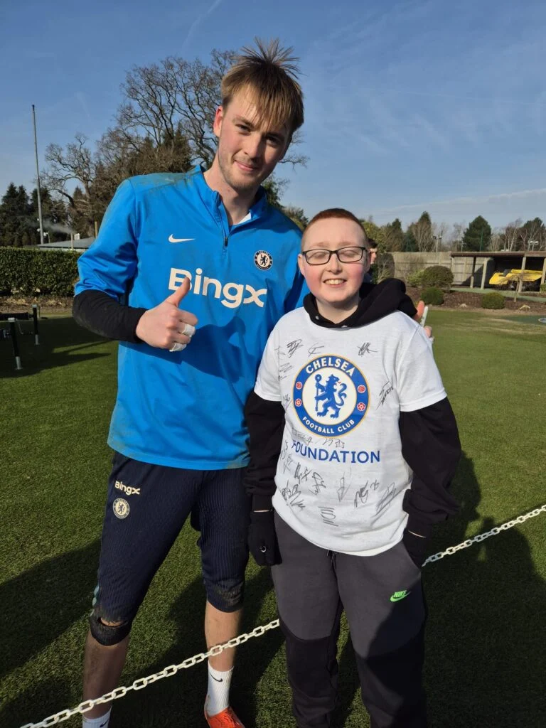 Young fan in a signed Chelsea Foundation shirt standing with a player in training kit