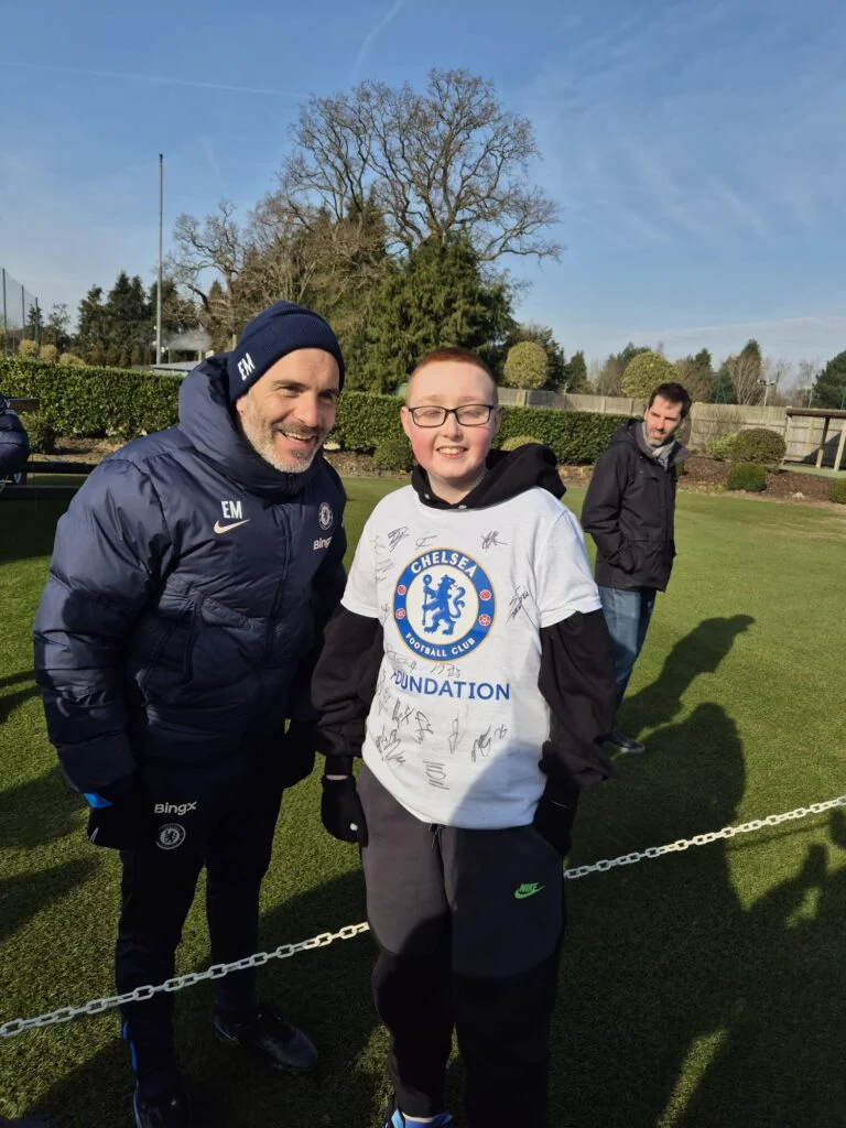 Young fan in a signed Chelsea shirt beside a member of coaching staff at the training ground